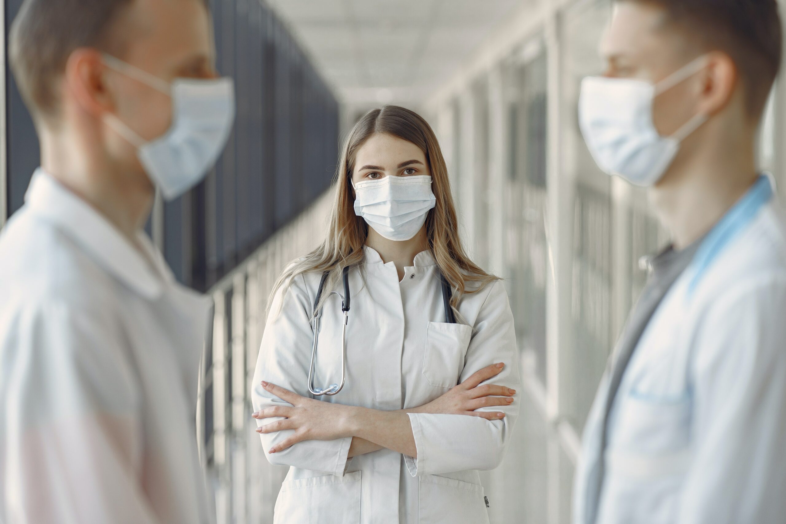 Zwei Mediziner stehen sich gegeüber und im Hintergrund eine weitere Medizinerin mittig guckt in die Kamera.